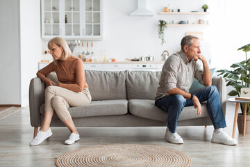 Older couple sits apart on a couch in a bright living room, showing tension and emotional distance....