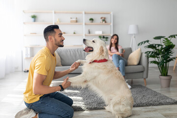 A young man interacts joyfully with a golden retriever in a bright living room decorated with plants. A woman is seated quietly on a couch, enjoying the moment.