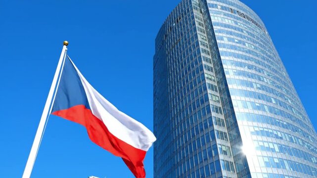 A white red and blue flag with a blue triangle flies on a pole in front of a modern glass skyscraper against a clear blue sky