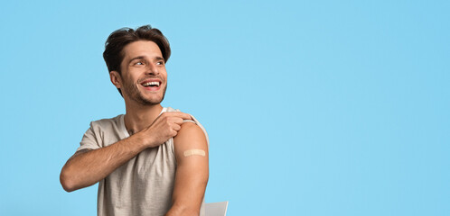 A young man with short hair is smiling while showing a bandage on his arm. He appears happy after getting a vaccination at a health clinic. The background is a light blue color.