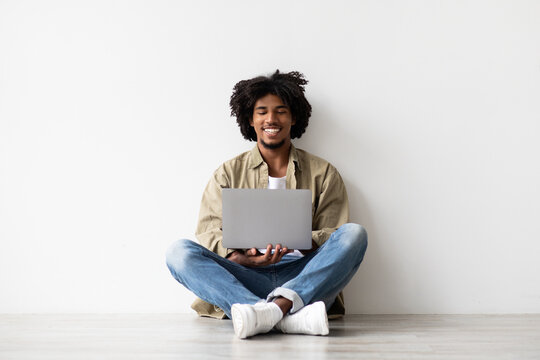 A smiling African American man sits comfortably on the floor near a white wall, typing on his laptop. He enjoys his distance job, radiating positivity and relaxation in his workspace.