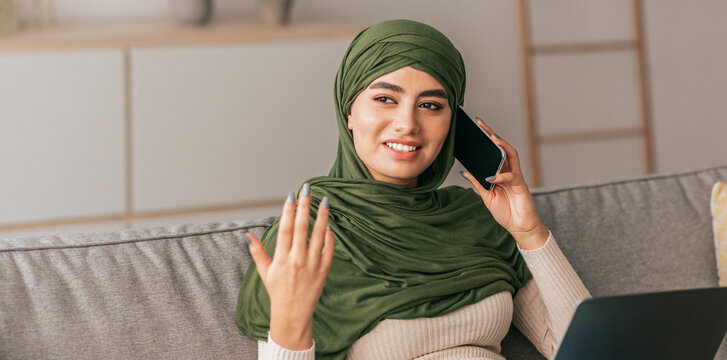 A young woman in a green hijab is engaged in a phone conversation while sitting comfortably on a sofa at home. She shows a friendly expression and gestures with her hand. - Powered by Adobe