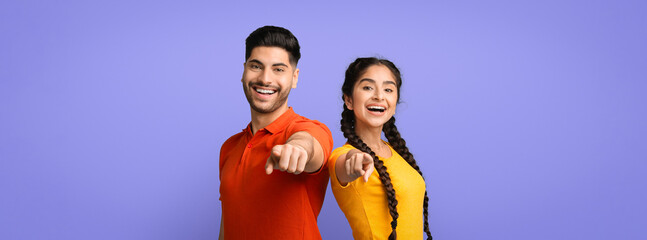 Two friends stand together against a light purple background, smiling widely and pointing at the...