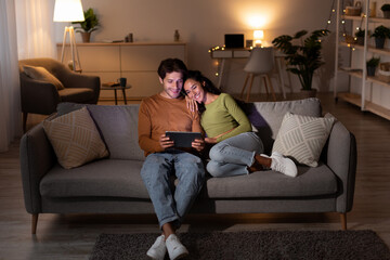 Obraz premium Young couple sits comfortably on their sofa in the evening, exploring the internet on a tablet.