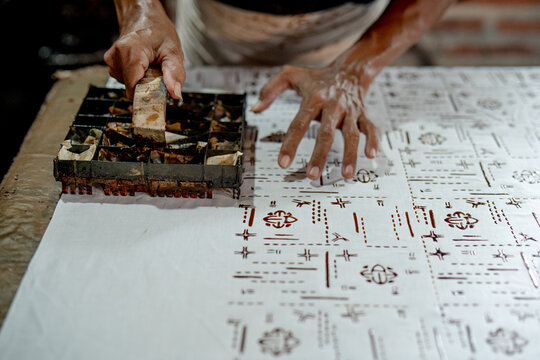 An artisan presses a metal batik stamp onto white fabric, creating traditional motifs in an Indonesian batik workshop. Batik Cap.