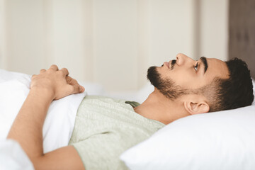 A young man with a beard lies in bed, staring at the ceiling. He appears thoughtful and contemplative, covered in a cozy blanket. Morning light softly fills the room.