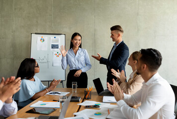 In an office boardroom, a diverse team greets a new Asian female colleague with smiles and applause. The supportive atmosphere showcases teamwork and collaboration among coworkers during the meeting.
