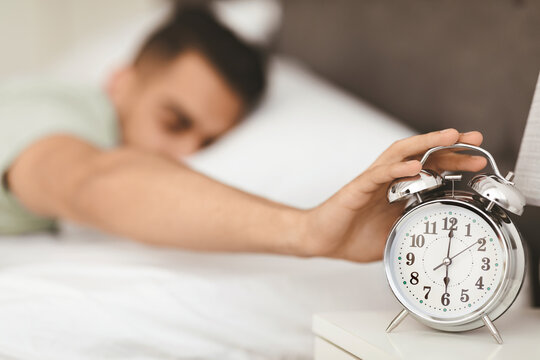 A man reaches out to turn off his alarm clock early in the morning. The cozy bedroom setting suggests a start to the day. Sunlight filters through the window, creating a peaceful atmosphere.