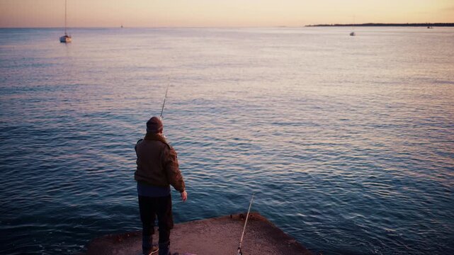 A man stands on a concrete pier while casting his fishing line into the sea at dawn