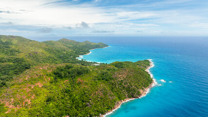Aerial view of green hills leading to quiet turquoise bays with white sandy beaches, tucked along a peaceful coastline. Praslin, Seychelles.