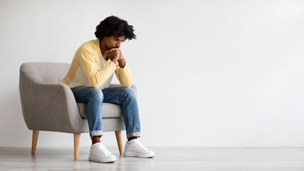 A thoughtful young African American man sits alone in an armchair, appearing lost in contemplation. He exhibits signs of mental distress in a quiet indoor setting, creating a scene of solitude.