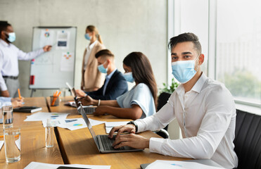 An Arab male employee wearing a face mask sits at a desk during a corporate business conference. He is using a laptop and looking at the camera while colleagues engage in a team discussion.