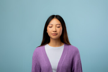 A young woman wearing a purple cardigan stands still with her eyes closed, embracing a moment of...