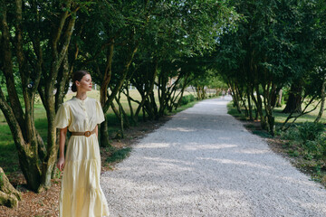 Fototapeta premium A woman in a flowing yellow dress walks along a gravel path bordered by trees, enjoying a peaceful outdoor park scene with dappled sunlight.