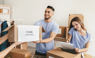 Two volunteers work together in a community center, sorting food donations for local families. One smiles while holding a box labeled food donations, and the other takes notes.