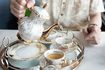 Close-up of a person pouring hot tea from a vintage floral porcelain teapot into a matching cup. Served on a golden tray for afternoon tea time.