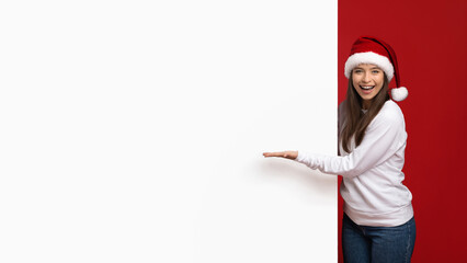 Obraz premium A young woman wearing a Santa hat and white sweater stands beside a bright red wall, smiling joyfully while presenting a blank space. The festive atmosphere enhances her cheerful spirit.