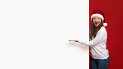 Fotobehang Ridders A young woman wearing a Santa hat and white sweater stands beside a bright red wall, smiling joyfully while presenting a blank space. The festive atmosphere enhances her cheerful spirit.  © Prostock-studio