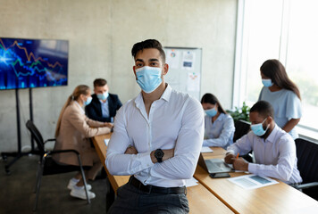Young middle eastern businessman stands confidently with folded arms in an office. Colleagues collaborate nearby while all are wearing medical masks amid the ongoing meeting.