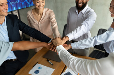 Diverse business professionals celebrate teamwork by stacking their hands together during a corporate meeting in a modern office. The atmosphere reflects unity and collaboration among coworkers.