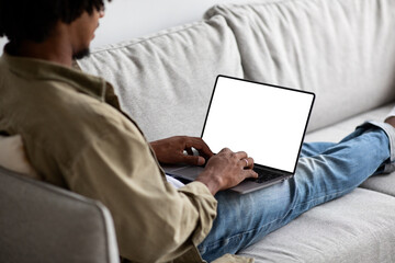 A black man is sitting on a couch at home, using a laptop with a blank white screen. He appears focused on his work, giving an over-the-shoulder view of him engaged with the device.