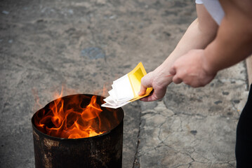 Hands holding joss paper (ghost money) over a burning fire in a metal bin. Traditional Chinese ritual for worshiping ancestors during Chinese New Year or Qingming Festival.