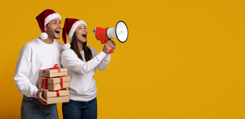 A cheerful couple wearing Santa hats stands against a bright yellow background. One person holds...