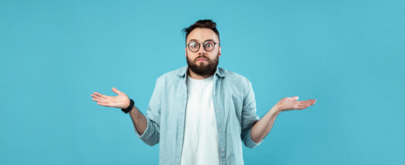 A man with glasses and a beard stands against a solid blue backdrop, shrugging his shoulders with a...