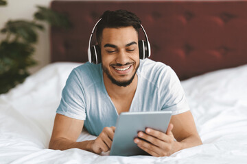 A young man lies comfortably on his bed, wearing headphones and smiling as he engages with a tablet. The cozy room features soft bedding and warm lighting, creating a peaceful atmosphere.