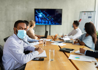 In a modern boardroom, diverse employees wearing face masks engage in a corporate meeting. They listen attentively to a presentation, discussing important business matters during the pandemic.