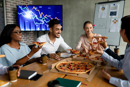 Colleagues from diverse backgrounds enjoy pizza together in the office during their lunch break. They are laughing and communicating while seated at a table filled with food and drinks.