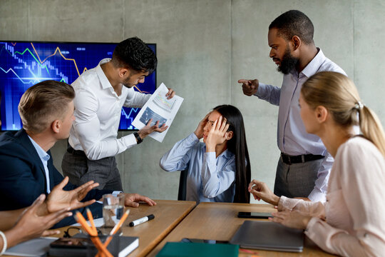In a modern office meeting, a multiethnic team argues over a failed project. An unhappy Asian employee faces criticism while coworkers express frustration and blame during the discussion.