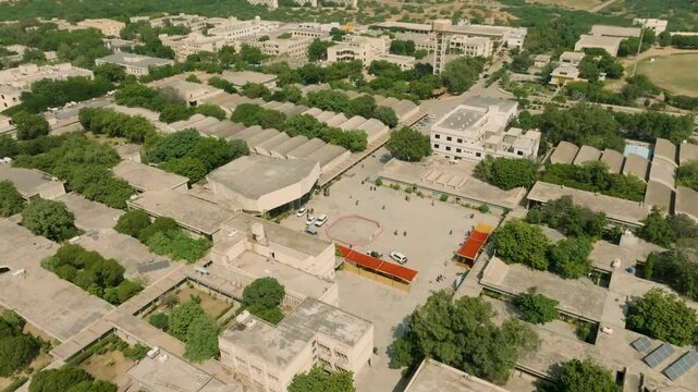 Drone descending and orbiting over the campus of NED University, highlighting beige buildings, green trees, and a central yard with people gathered on a sunny afternoon.