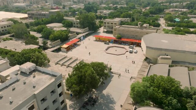 Drone receding and ascending over bustling central open yard of NED University, showing people walking, trees, campus buildings, and the illuminated Karachi city skyline in the background.
