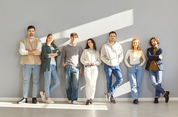 Obraz premium Group of fashion models posing in casual clothing. Seven happy young people wearing comfortable informal clothing standing by a gray wall in sunlight. Fashion concept