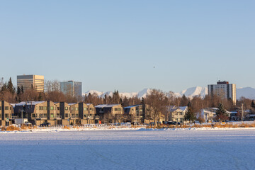 Anchorage Residential District with Downtown Skyline. Peaceful suburban scene in Anchorage, Alaska, showing houses, trees, snow, and the urban skyline set against mountain peaks.