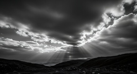 Dramatic sun rays pierce through dark stormy clouds over a rugged mountainous landscape in black and white
