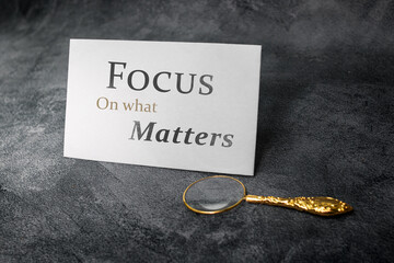 A motivational card with the message “Focus on what Matters” stands on a dark textured surface, accompanied by an elegant gold magnifying glass, symbolizing clarity, priorities, and mindful direction.