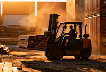 Silhouetted worker operating a forklift, carrying a heavy load of lumber planks at sunset in a bustling outdoor freight yard or container port, with dust and steam in the warm sunlight