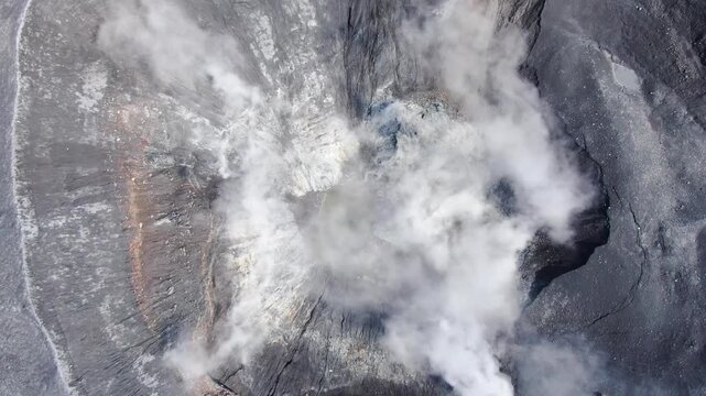Dramatic 4K shot of the drone's orbit with a smoking volcanic crater, with thick white smoke, showing unbridled power and a desolate volcanic landscape. Kuril Islands.