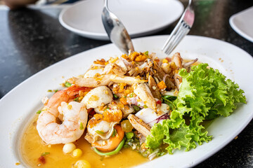 Close-up of fork and spoon lifting pieces of spicy seafood salad with shrimp and squid from a saucy white plate.