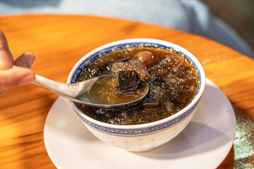 A spoon lifting a piece of black grass jelly from a bowl of ice and syrup on a wooden table.