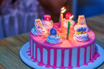 A pink birthday cake decorated with cream colorful toppings and lit candles on a wooden table.