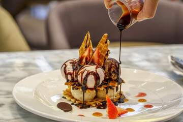 Hand pouring chocolate sauce onto a waffle dessert topped with ice cream and strawberries.