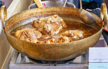 Tender stewed pork leg boiling in brown gravy sauce in large brass pan at street market.