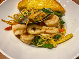 Spicy seafood spaghetti stir-fry featuring shrimp, squid, mussel, and herbs, served with a crispy slice of garlic bread on a white plate.