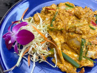 A rich and creamy stir-fried crab dish with yellow curry powder, egg, and vegetables, garnished with a purple orchid on a blue plate.