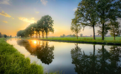 Obraz premium A picturesque sunset beautifully reflecting on a tranquil river, surrounded by lush, vibrant trees and grass