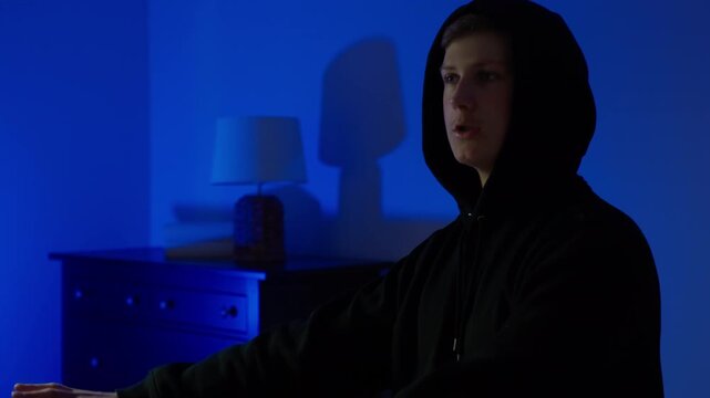 Hooded young man stands inside dark house interior, hands raised in gesture of confusion or surrender, harsh red and blue alarm light emphasizes emotional stress and uncertainty during crime.
