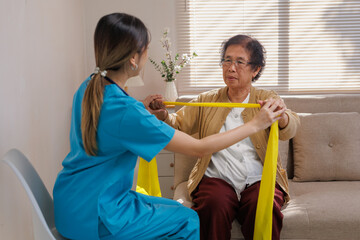 Female nurse assisting an elder woman with arm exercises using a yellow resistance band for rehabilitation at home